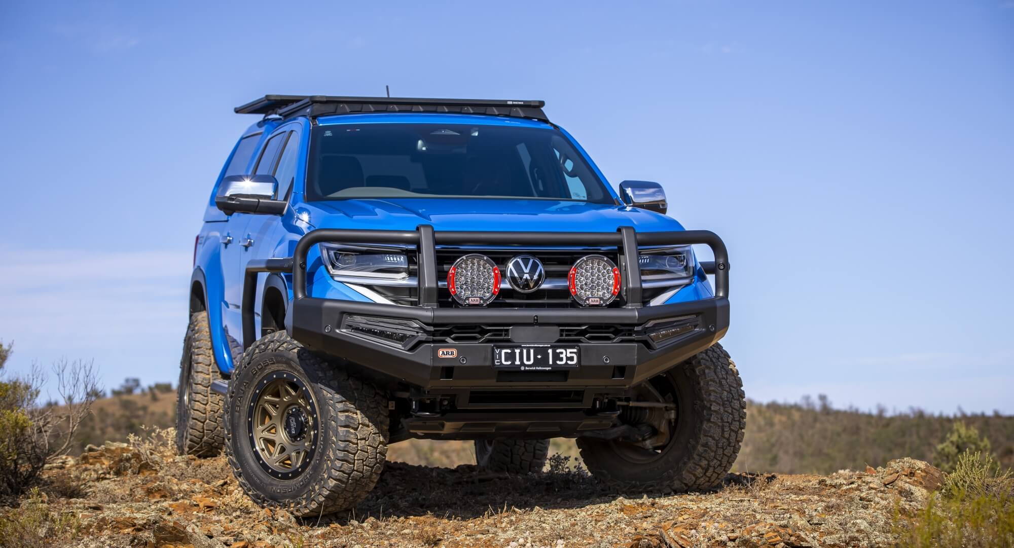 ARB Blue Volkswagen Amarok sitting on a slight angle on the top of a grassy hill. Showcasing Black Summit MKII Bull Bar and Intensity Solis 36 Driving Lights combination set up with red bezels