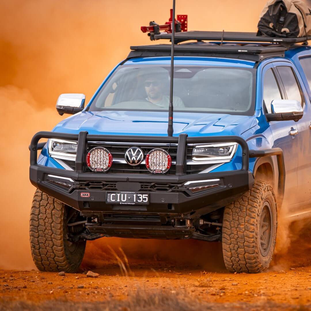 ARB Blue Volkswagen Amarok driving in and orange dusty area. Amarok showing Black Summit MKII Bull Bar, Intensity Solis 36 Driving Lights and a base rack on the roof of the vehicle.