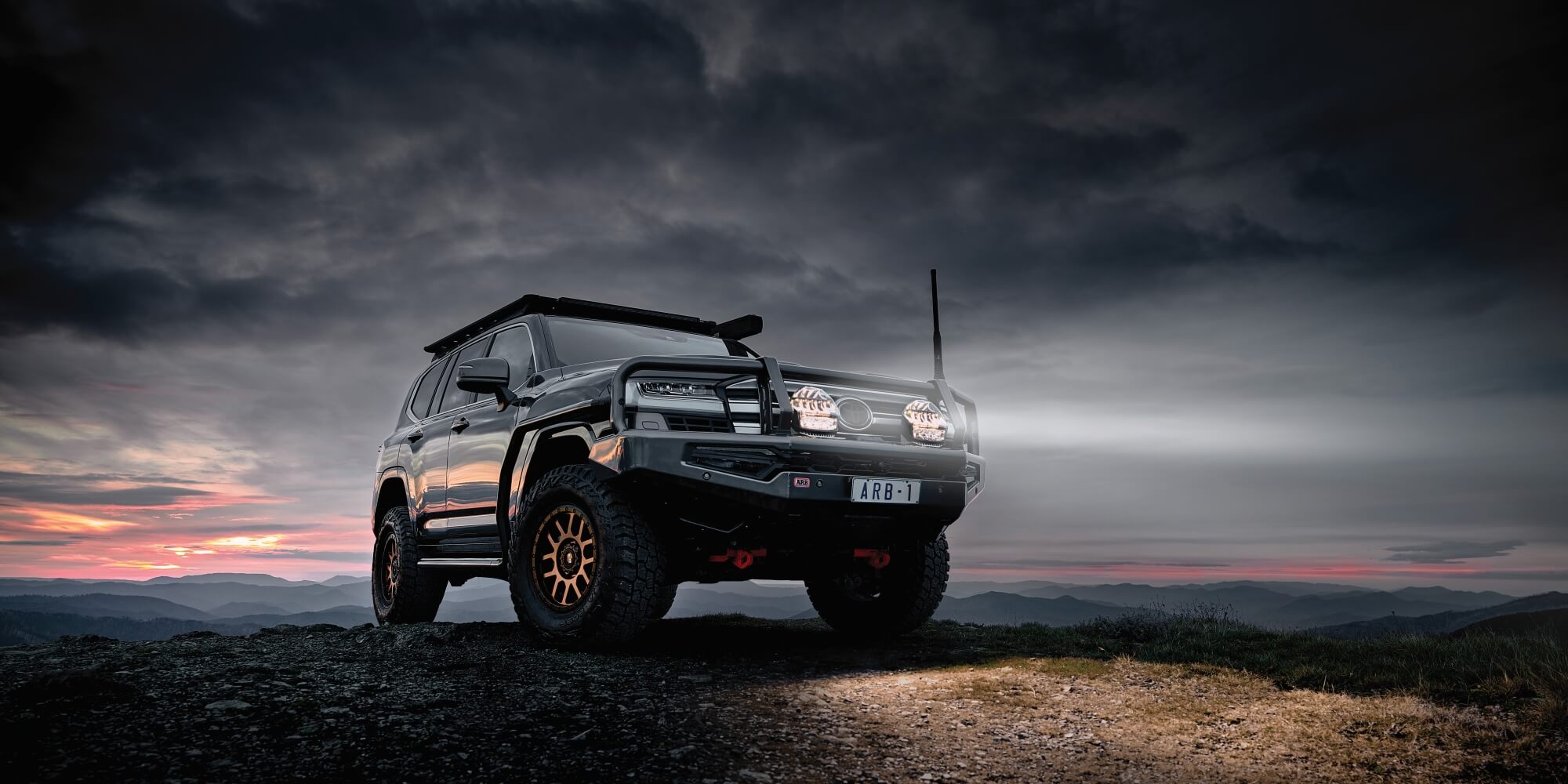 ARB Toyota 300 series ontop of a hill with the sunset behind the car showing the Intensity IQ driving lights mounted on top of a black Summit MKII Bull Bar